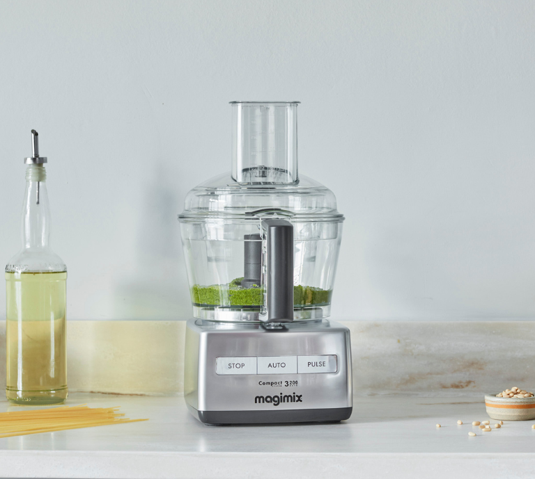 Photograph of a Magimix CS 3200 Compact Food Processor, featuring ingredients for a pesto including olive oil, pine nuts and basil