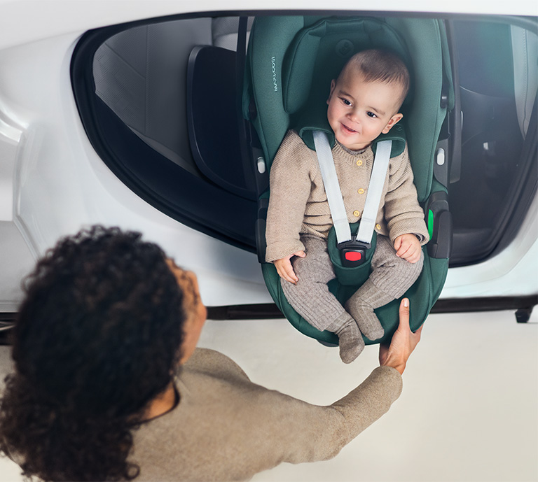 Green maxi-cosi car seat showing a baby in the product and a mother sliding the baby out from the car