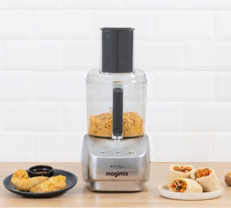 Photograph of a Magimix Mini Plus Food Processor in a kitchen with breadcrumbs in the main bowl and ingredients on the side like lime, carrots and courgette.