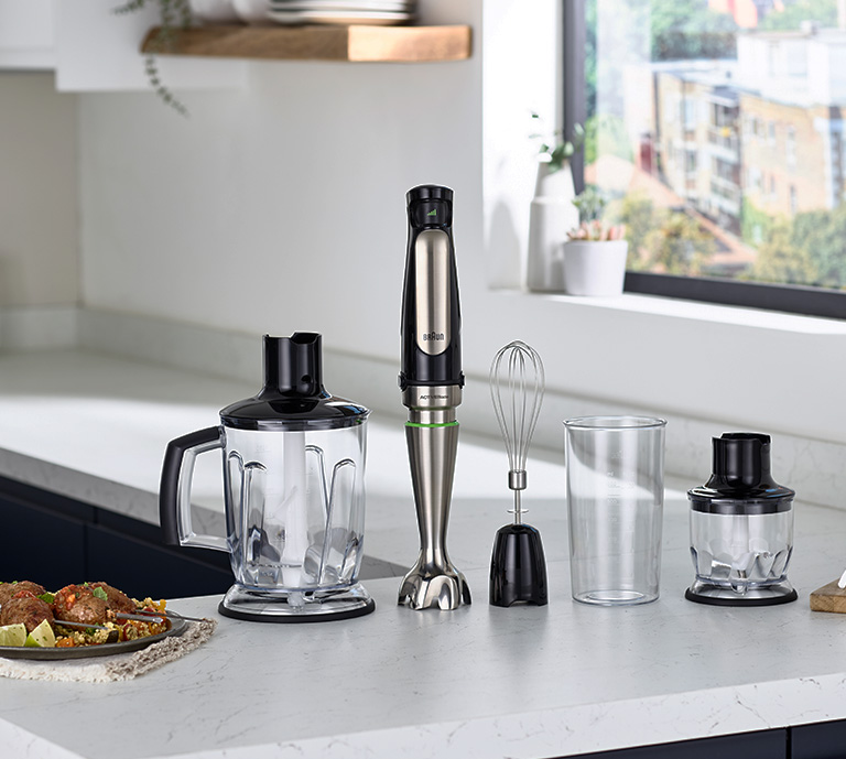 Braun hand blender sat on a kitchen counter top. Plate of food to the left of the image.