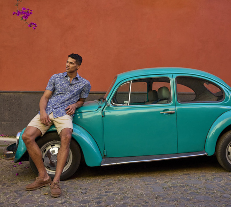 An image of a man wearing a blue shirt and chino shorts sat on an old teal car on a street in Mexico. This is advertising Crew Clothing's shirt collection and includes a link to go through to the page