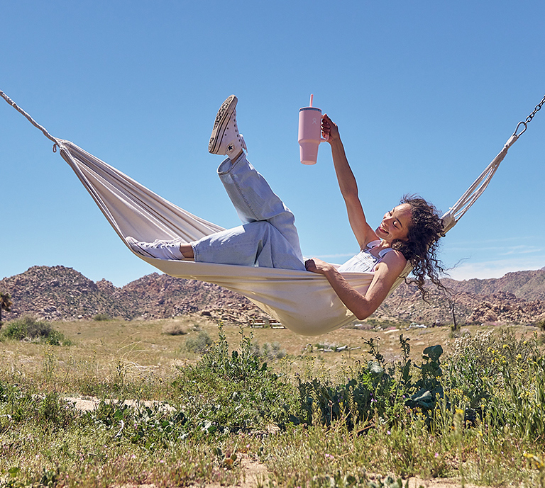 Hydro Flask new All Around Travel Tumbler Lifestyle imagery of young woman in a hammock outside holding Travel Tumbler in Trilium color